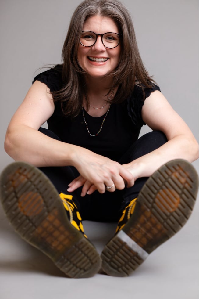 Brie’Anne sitting cross-legged on the floor, wearing dark clothes and bold black Doc Martens with yellow smiley faces, soles visible, in a relaxed pose with a smile.